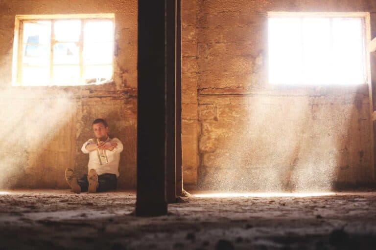 A person sits on the floor in a dusty room with sunlight streaming through two windows in a rough, worn wall.