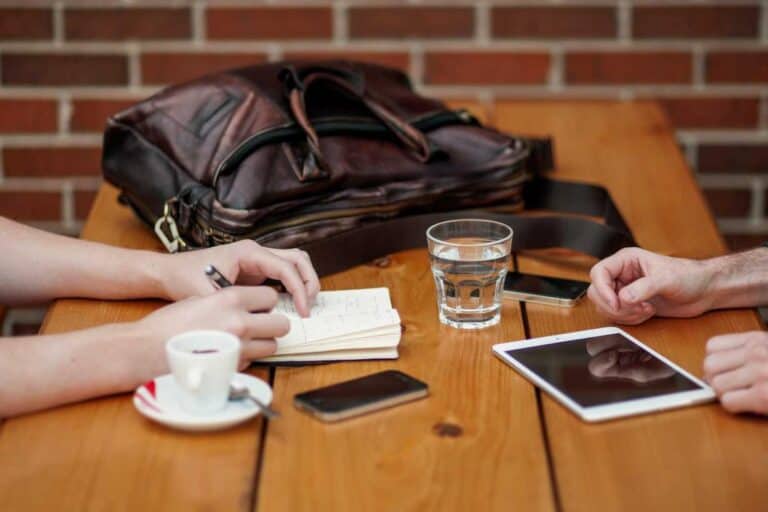 Two people sit at a wooden table with a notebook, cup, glass of water, phones, tablet, and a leather bag nearby.