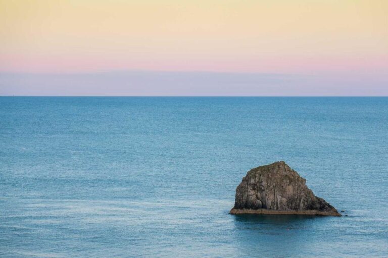 A large rock sits in calm blue ocean water under a pastel-colored sky at sunset or sunrise.
