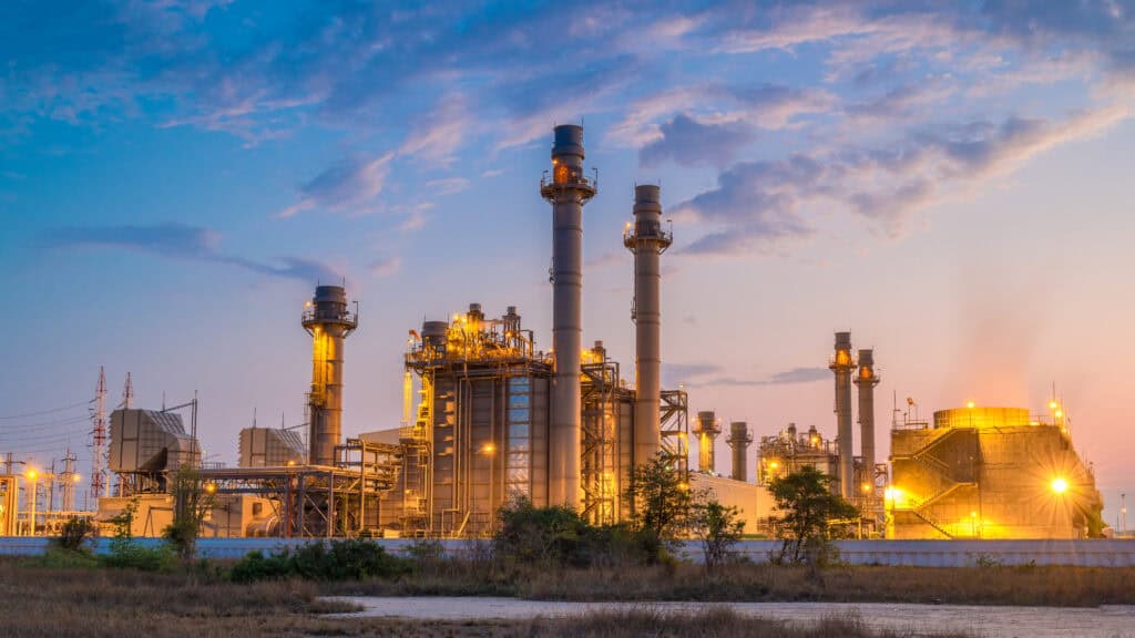 A large industrial power plant with tall chimneys and lit structures at dusk, with a cloudy sky in the background.